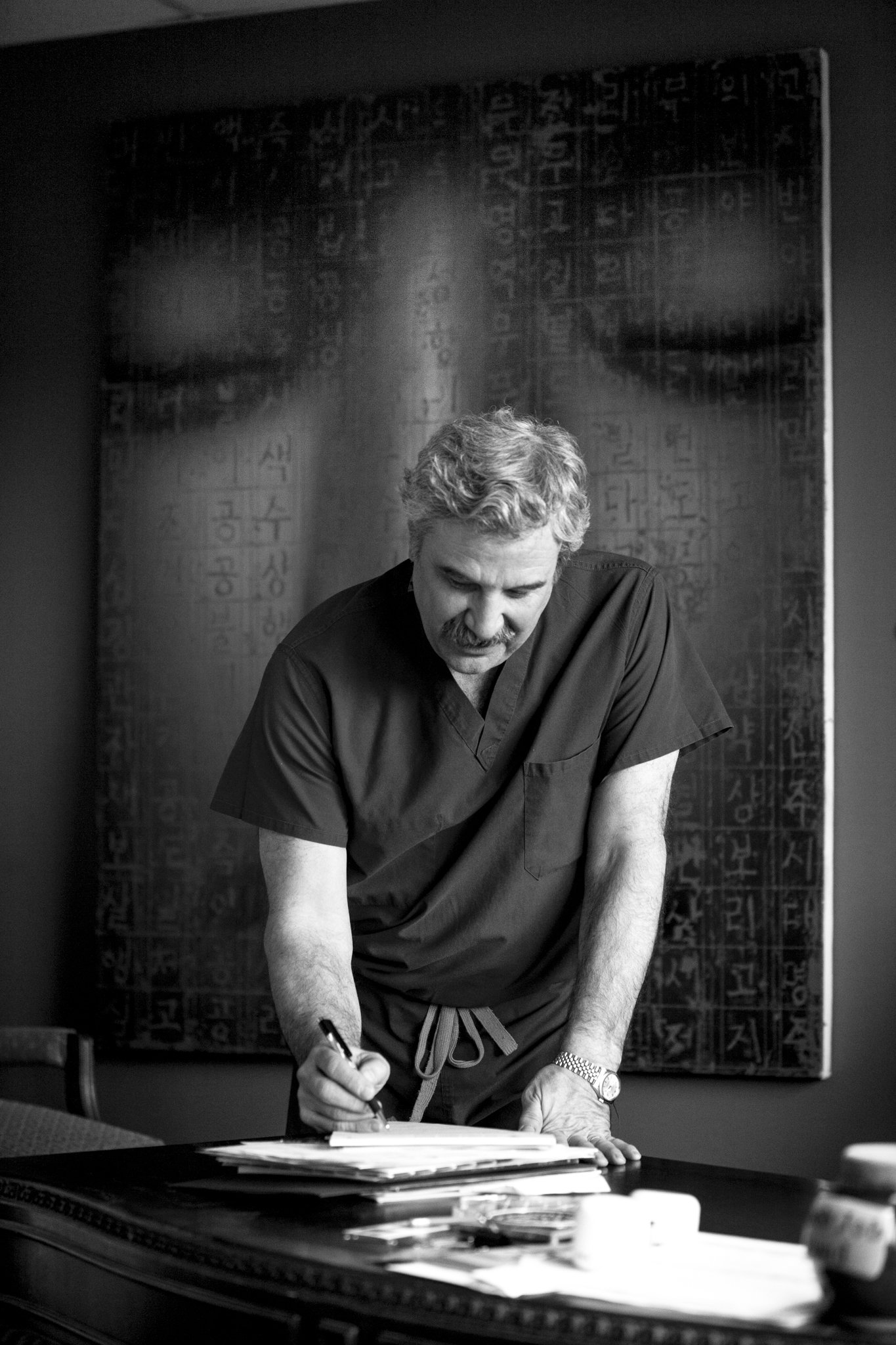 A man with gray hair and a mustache signing a document on a table, with stacks of papers and a pen in his hand, in front of a dark wall with abstract artwork in black and white.