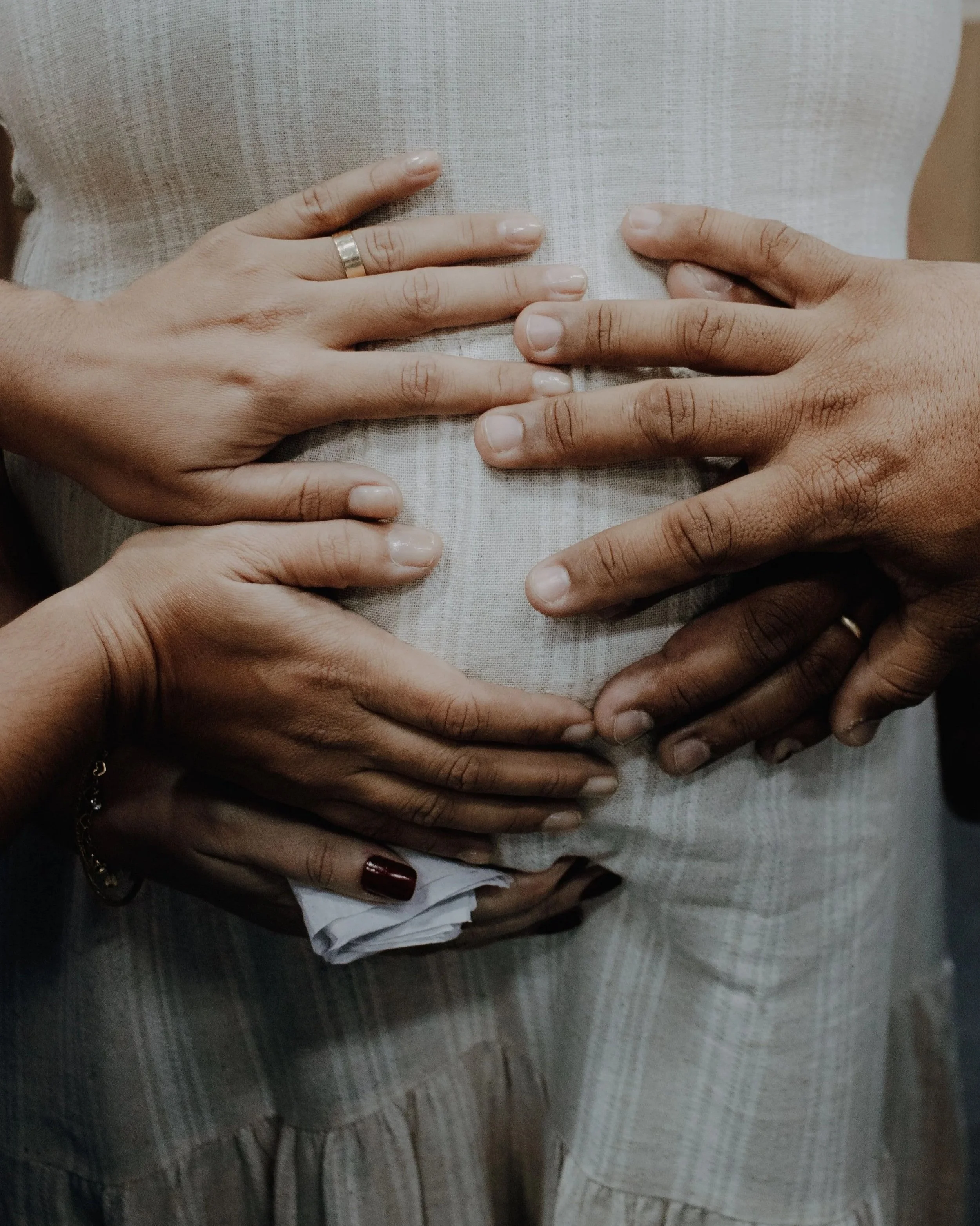 Multiple hands placed on a person's abdomen, forming a heart shape, with some hands wearing rings.