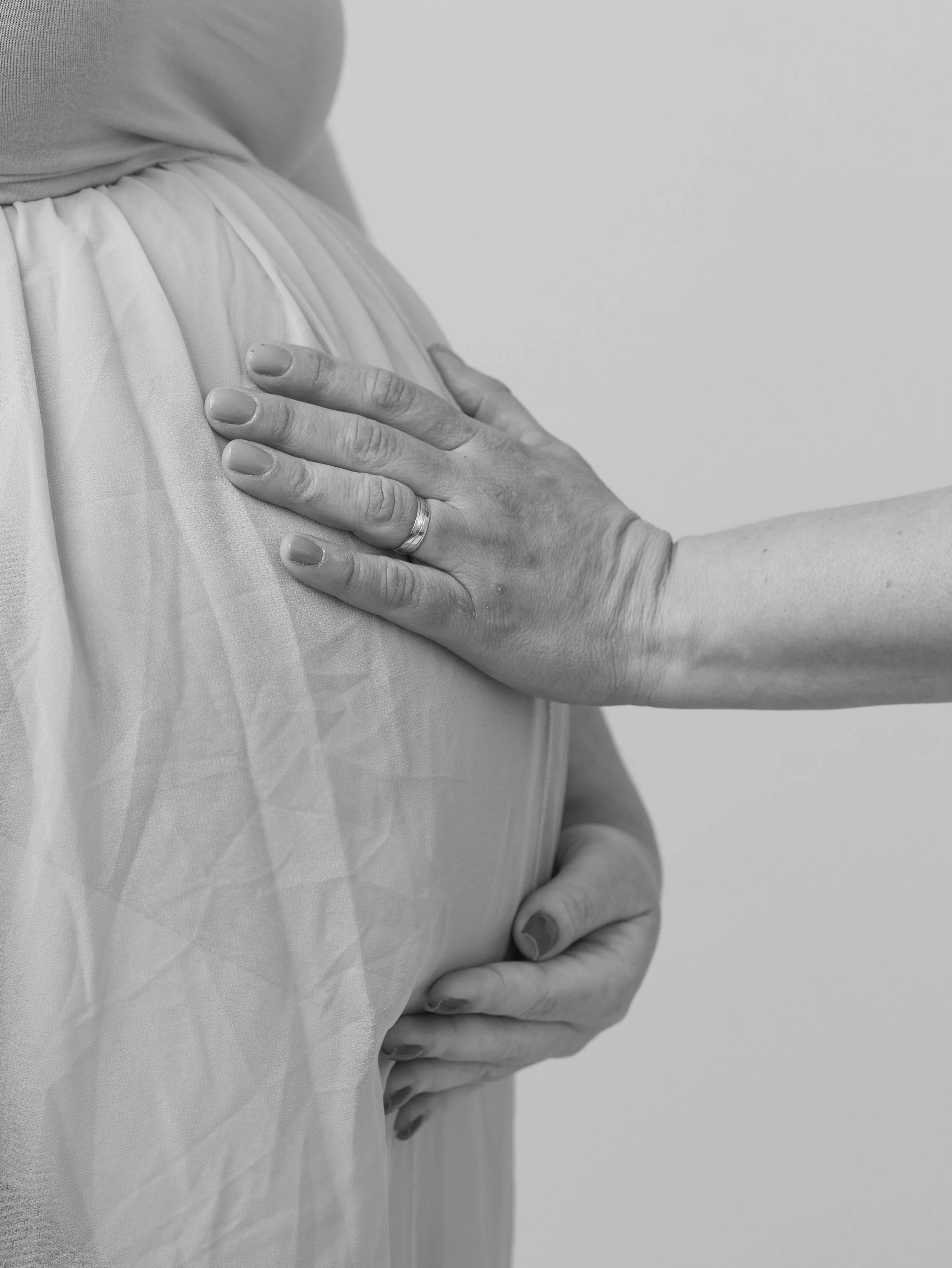 Close-up of a woman's hand on her pregnant belly, wearing a ring on her ring finger, with her fingernails painted. The woman is dressed in a light-colored, pleated fabric garment and the background is plain and softly blurred.