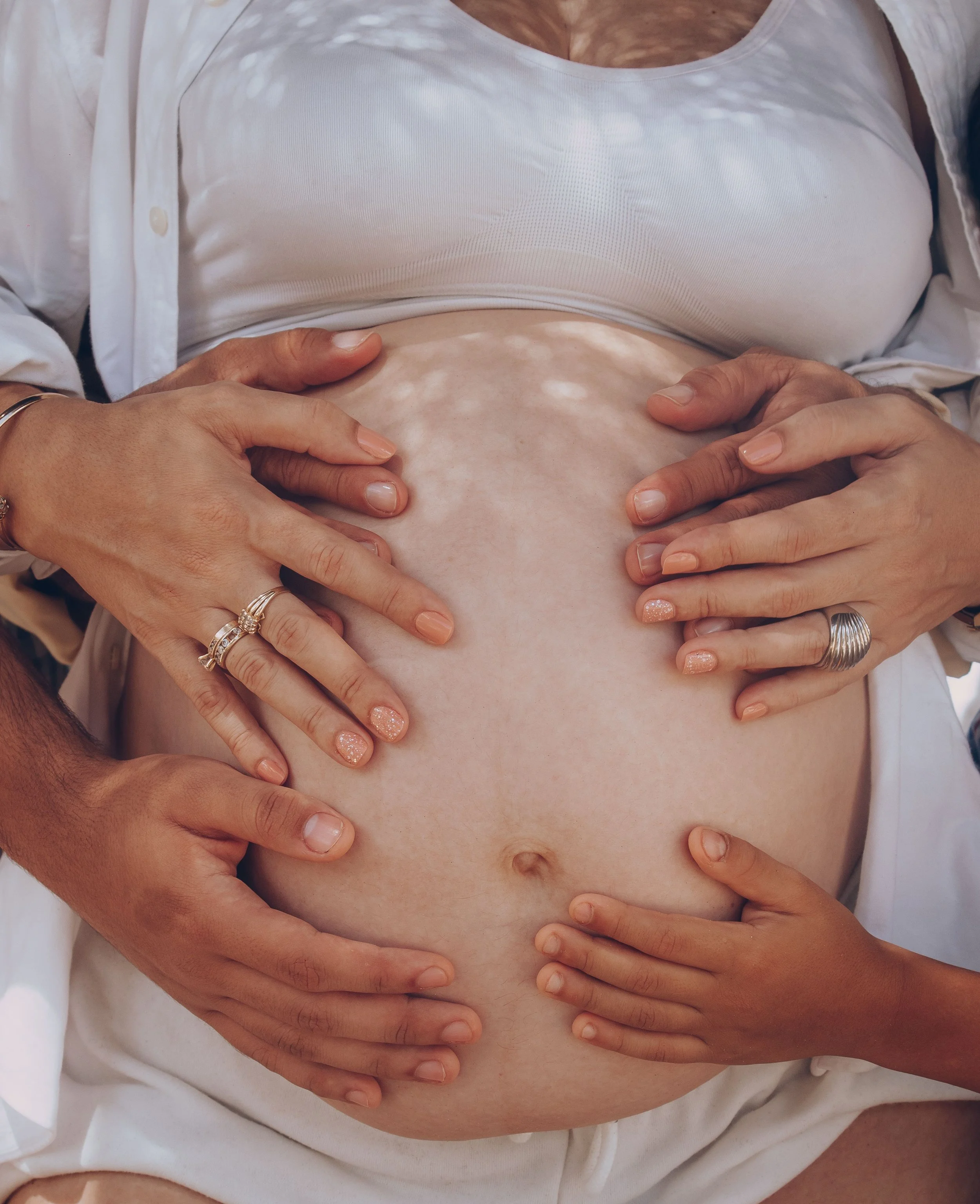 Close-up of a pregnant woman with her hands on her bare, rounded belly, wearing a white tank top and an unbuttoned shirt.