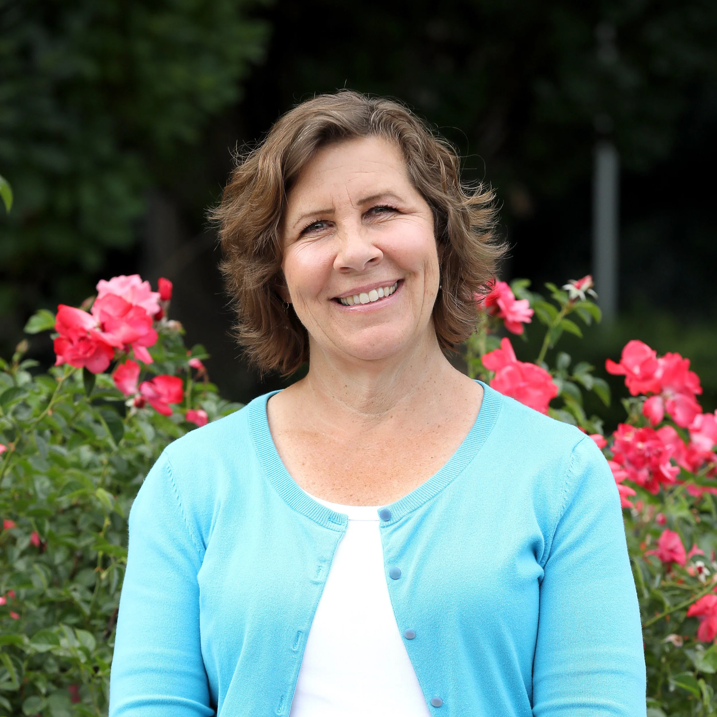 Portrait of a smiling middle-aged woman with brown hair, wearing a light blue cardigan over a white top, standing outdoors with pink flowers and greenery in the background.