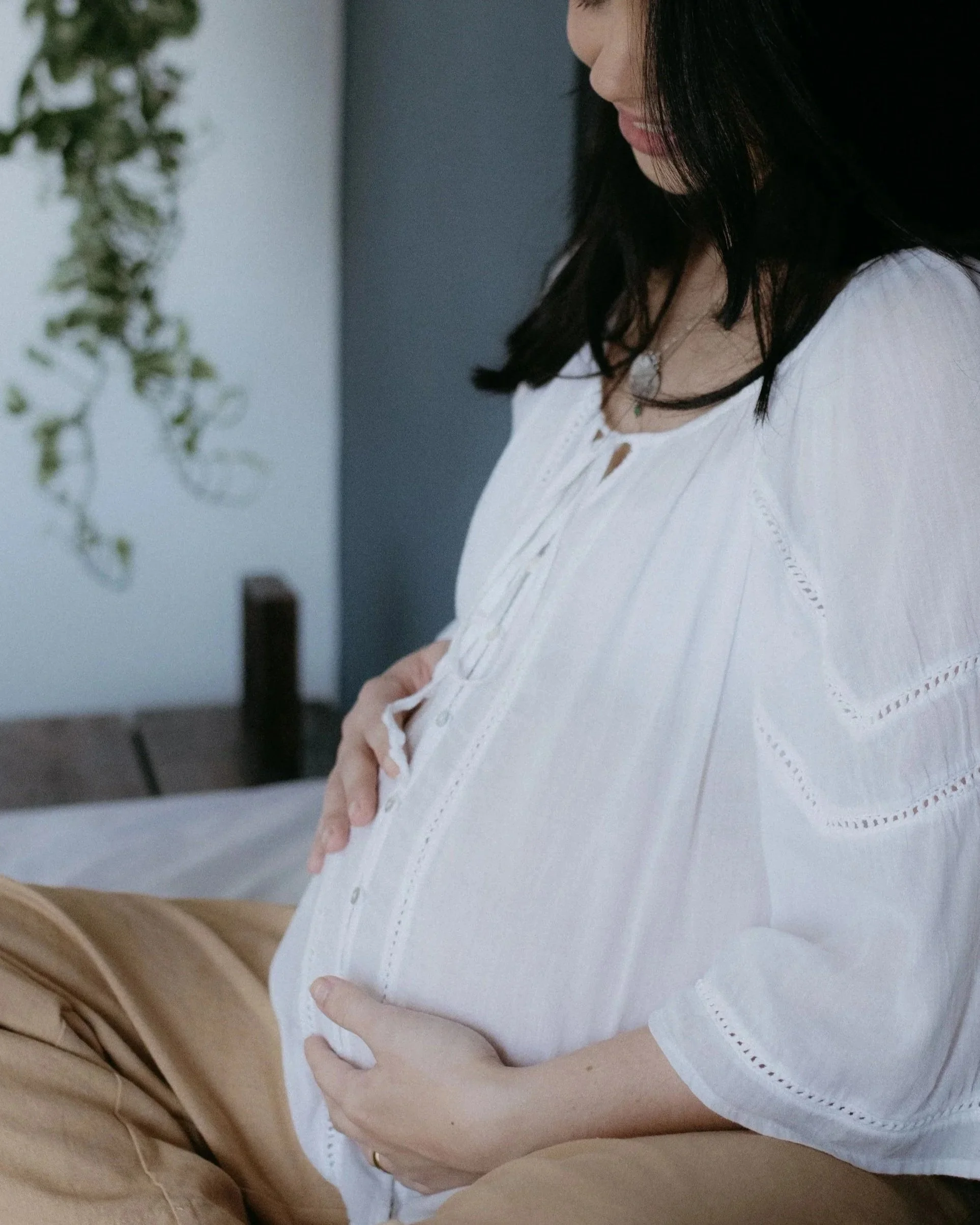 A woman with dark hair wearing a white blouse standing close to a man with tan pants. The woman is smiling and touching her stomach, possibly pregnant, while the man is sitting and holding her waist.