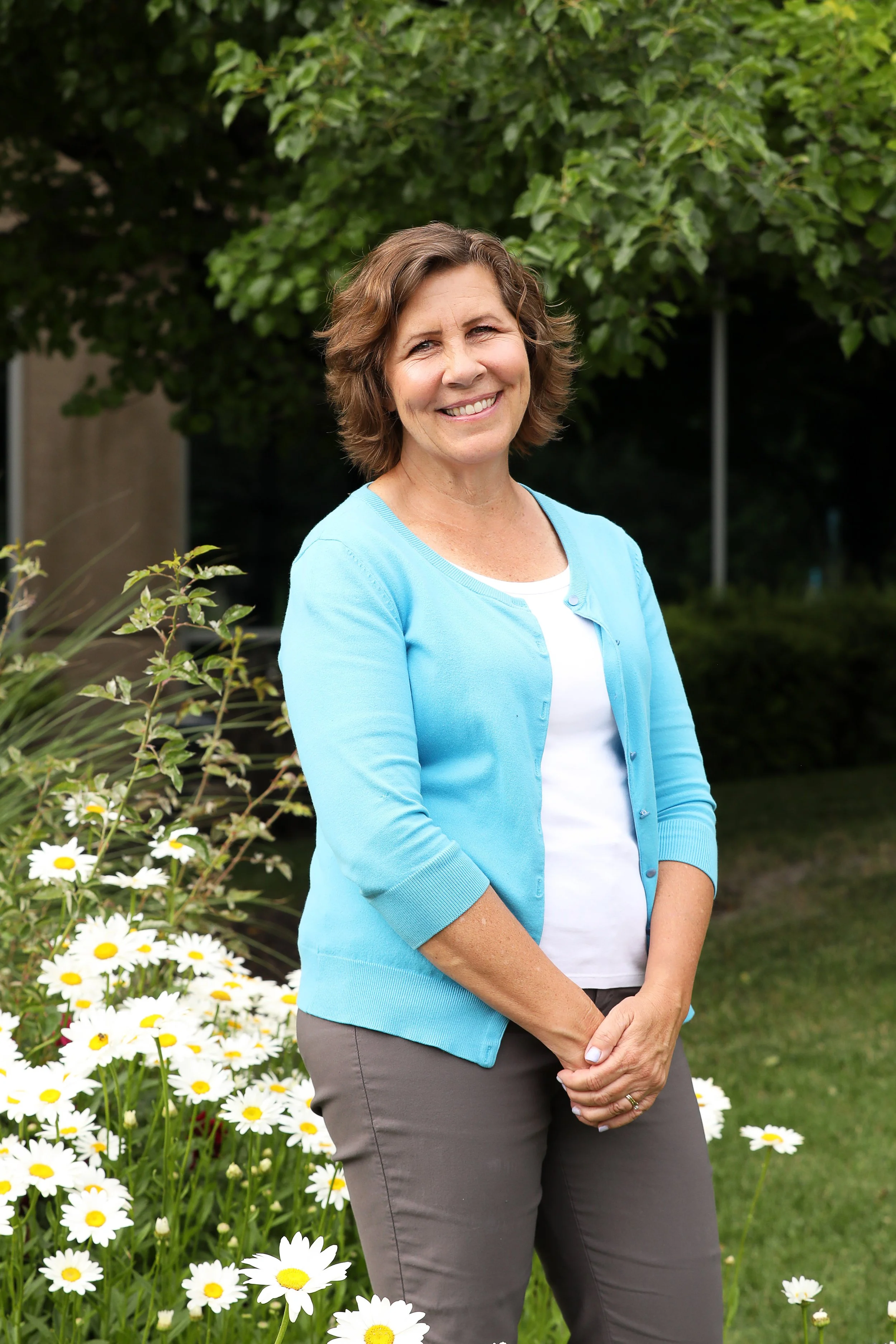 A middle-aged woman standing outdoors in front of a green leafy tree and a bed of white daisies with yellow centers, smiling at the camera, wearing a light blue cardigan over a white shirt and gray pants.