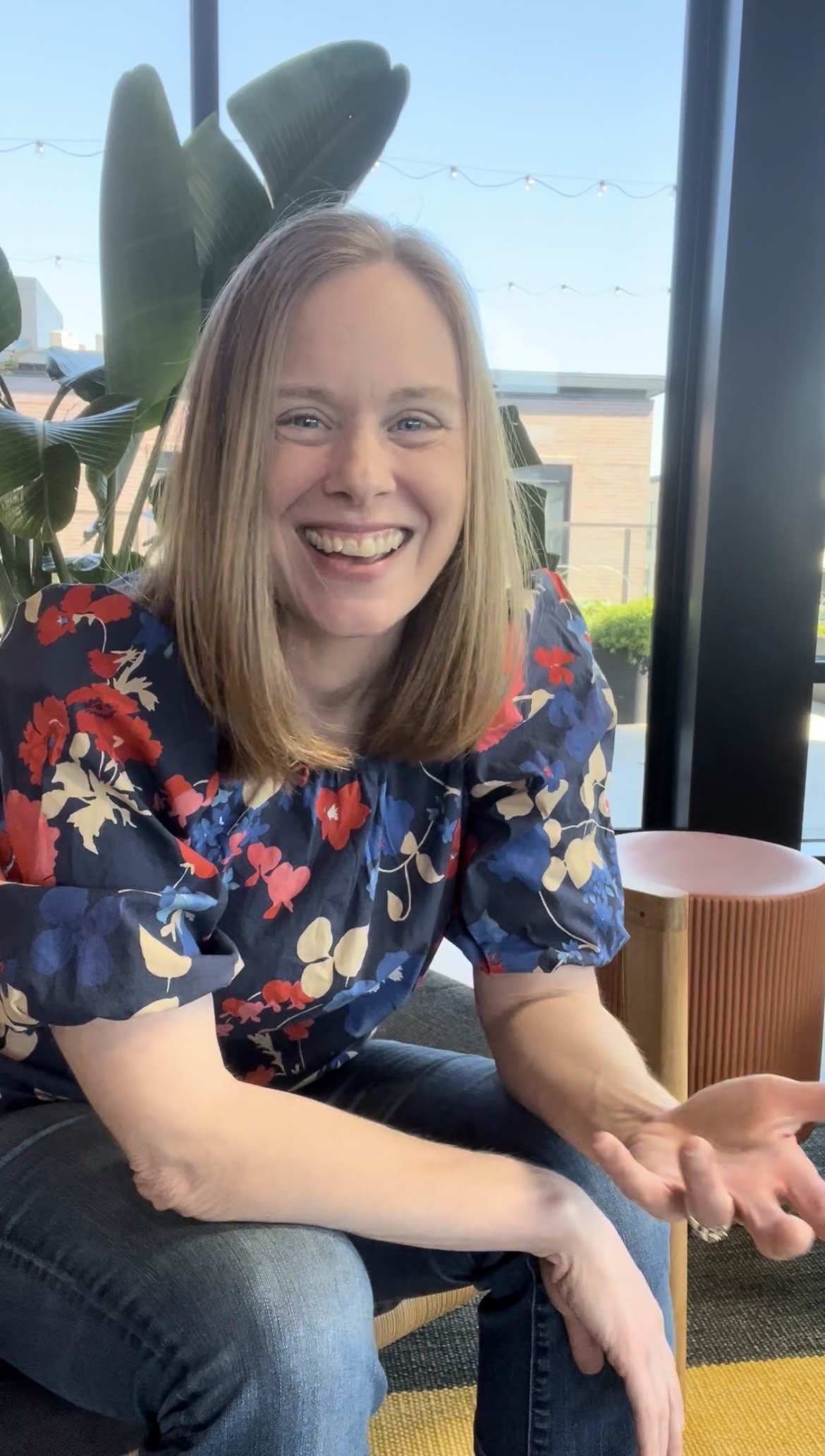 A smiling woman with blonde hair wearing a floral blouse, sitting indoors near a large window with plants and string lights visible outside.