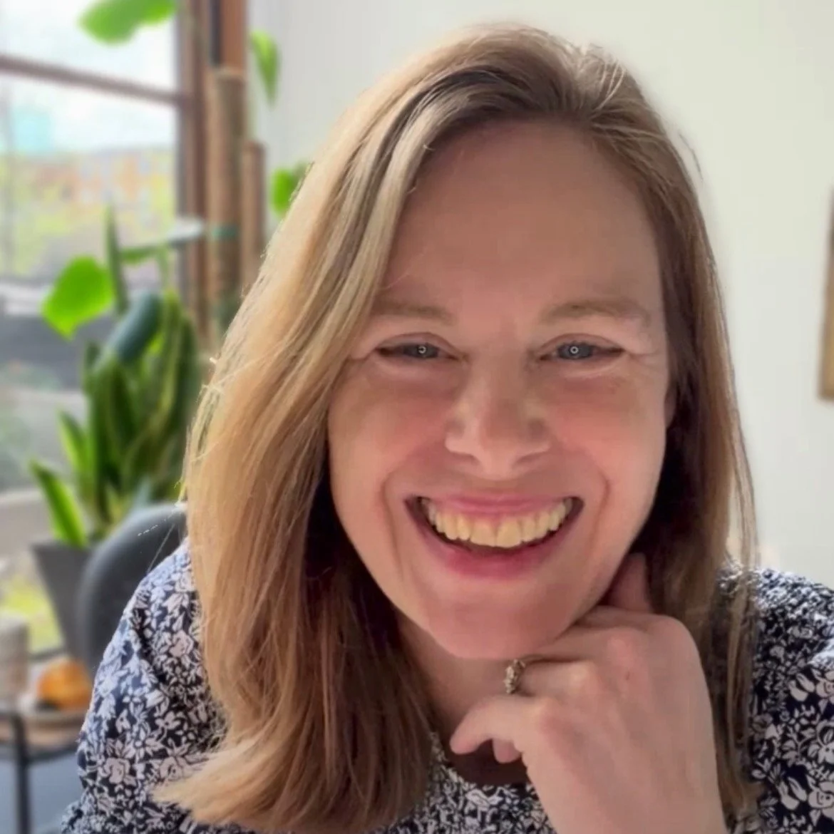 A woman with light brown hair smiling and resting her chin on her hand indoors, with a plant and window in the background.