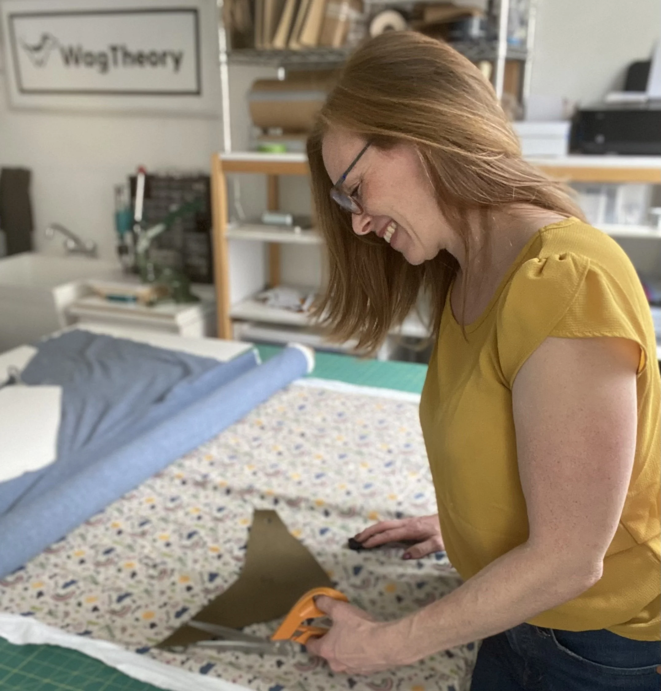 Person in yellow shirt sitting on floor with hands on the ground, smiling and leaning forward, in a room with shelves and a sign that reads 'WagTheory' in the background.