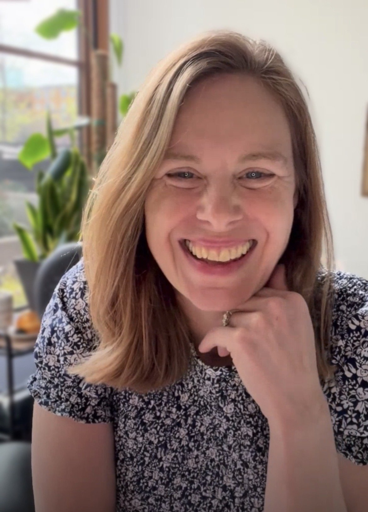 A woman with shoulder-length light brown hair smiling, resting her chin on her right hand, sitting indoors near a window with green plants in the background.