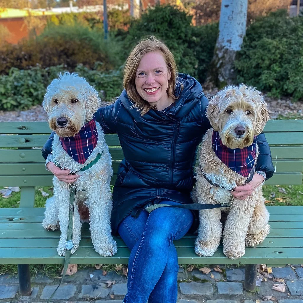 A woman sitting on a green park bench outdoors, smiling, with two large, fluffy, light-colored dogs with bandanas around their necks on either side. The background features trees and shrubs.