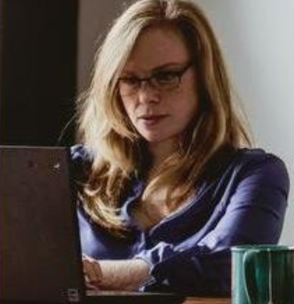 A woman with blonde hair, glasses, and dark lipstick sitting at a table with a laptop in front of her, wearing a dark blue shirt.