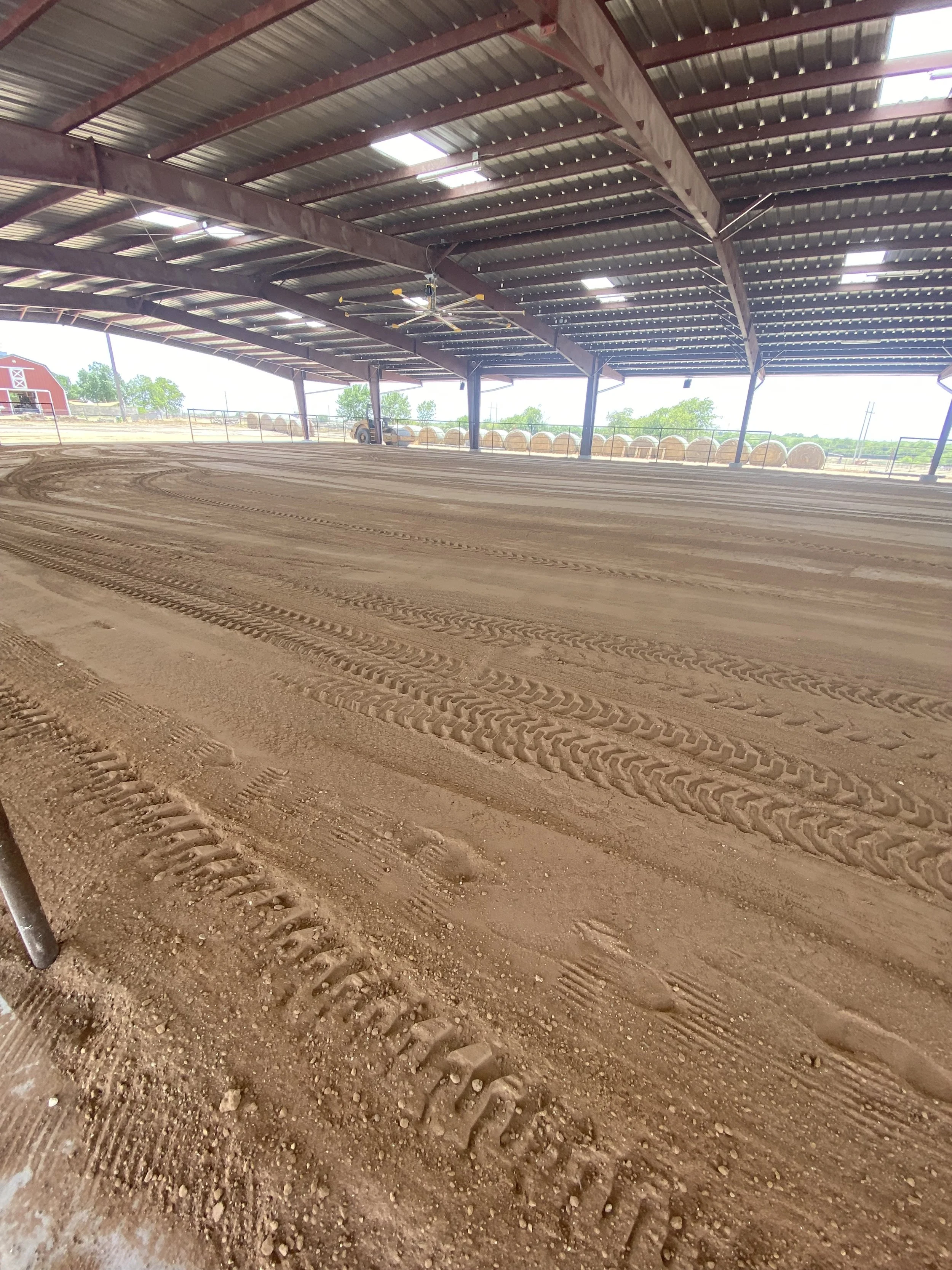 An indoor arena with a dirt floor, tire tracks, and a covered metal roof. Hay bales are visible outside, along with a red barn and green trees.
