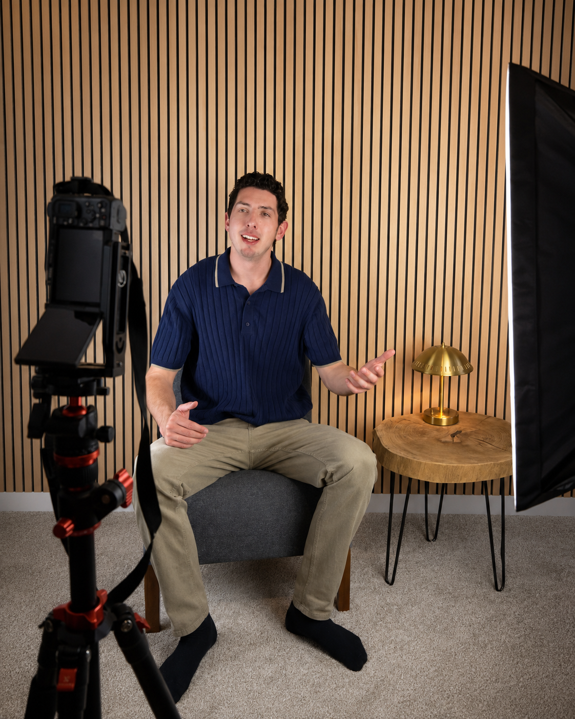 A man sitting on a gray ottoman in a recording studio, speaking or explaining, with professional camera and lighting setup, a wooden wall background, and a small wooden side table with a gold desk lamp.