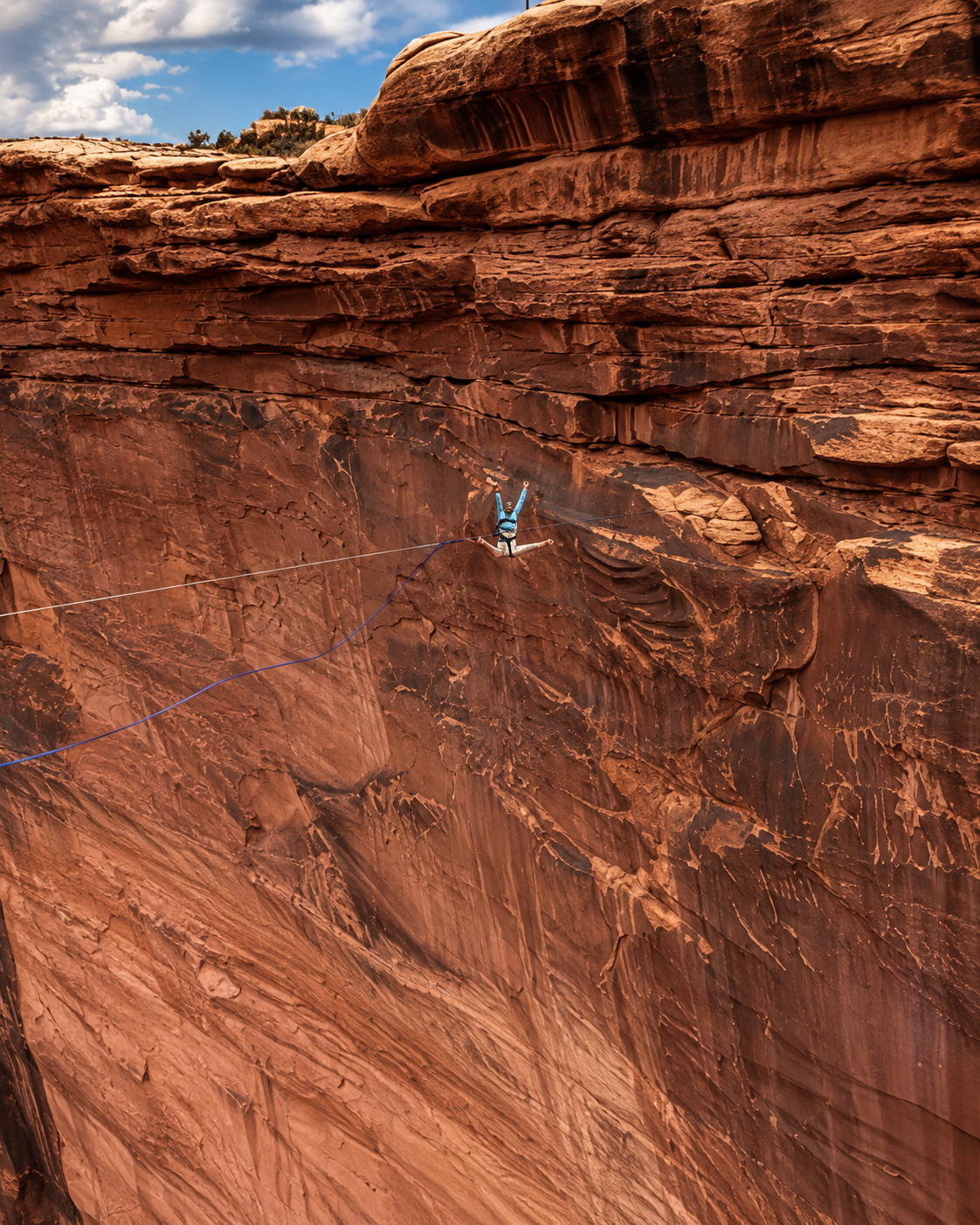 A person doing a handstand on a slackline stretched across a red rock canyon, with layered rock formations and a cloudy blue sky in the background.