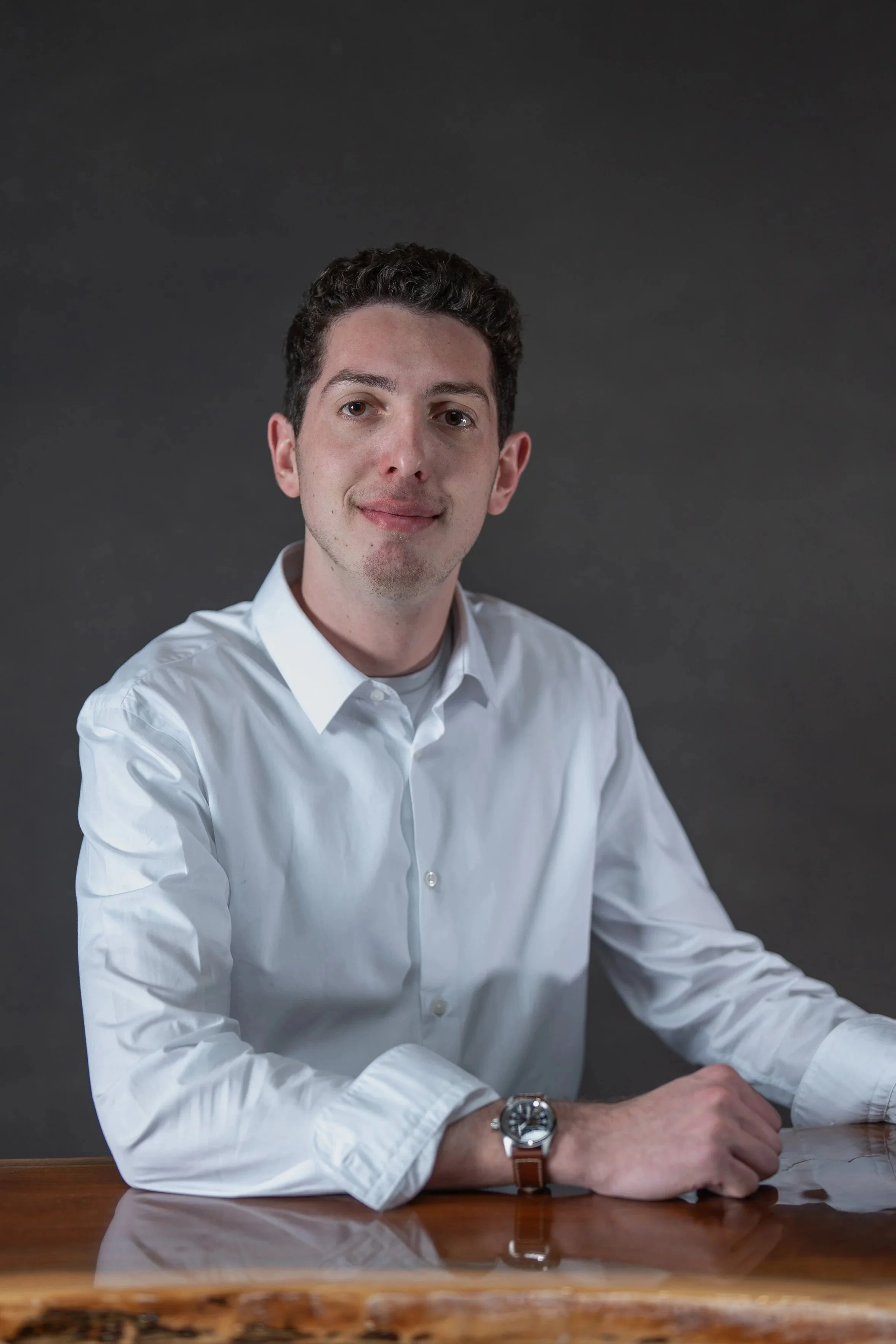 A man with short dark curly hair, wearing a white dress shirt, sitting at a wooden table, against a dark background.