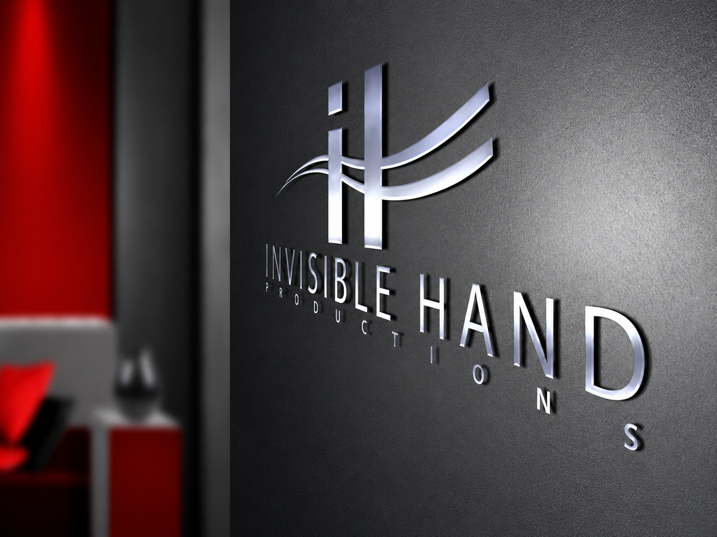 A wall sign displays the logo and name of 'Invisible Hand Production' with a sleek, modern design. The logo consists of stylized lines and the company's name is in bold, metallic lettering on a dark textured background. The left side of the image shows a blurred red and gray interior space.