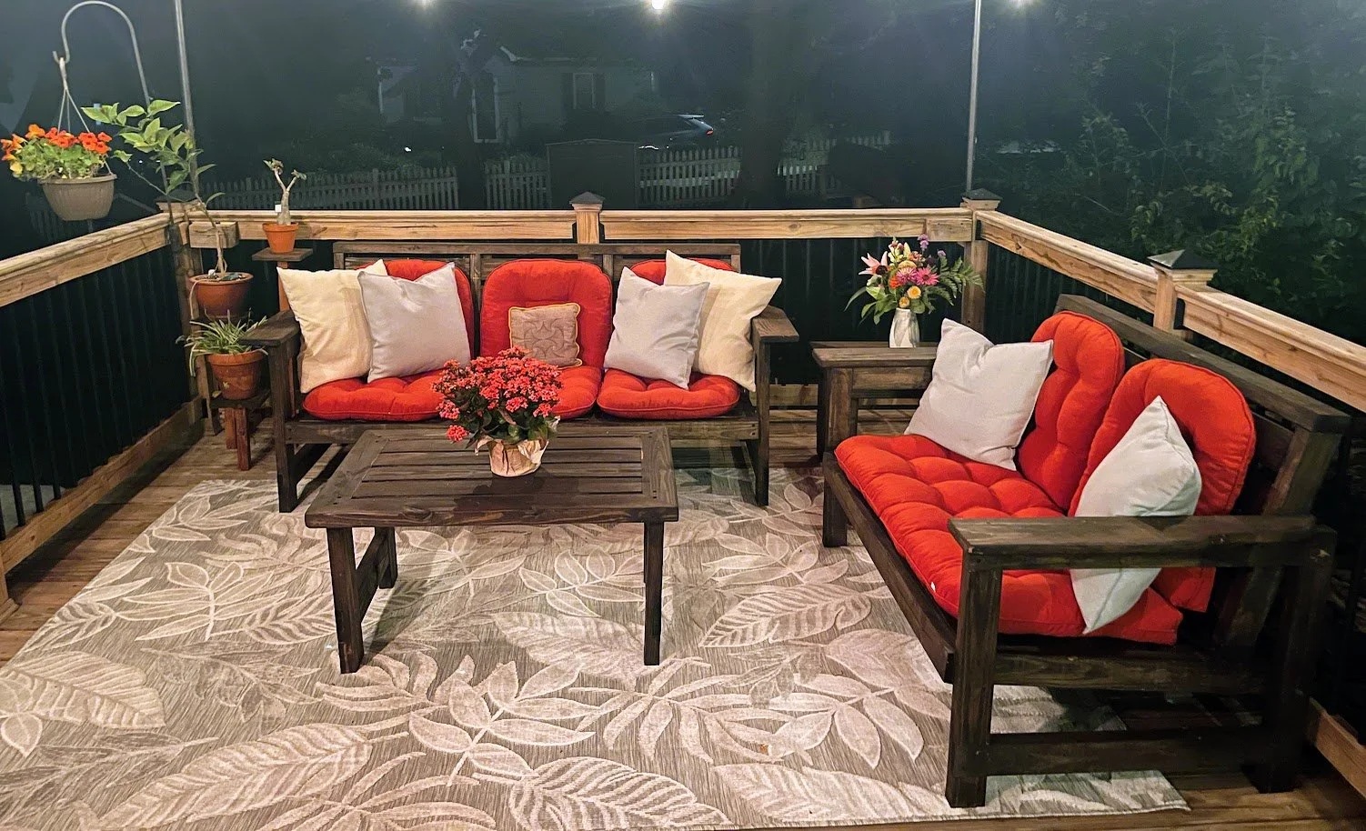 A cozy outdoor patio with wooden furniture, including a couch and chairs with red cushions and white pillows. There are potted plants on side tables and hanging baskets, a large floral arrangement, a potted pink flower, and an area rug with a tropical leaf pattern. The setting is illuminated at night with a dark background and a fence surrounding the patio.