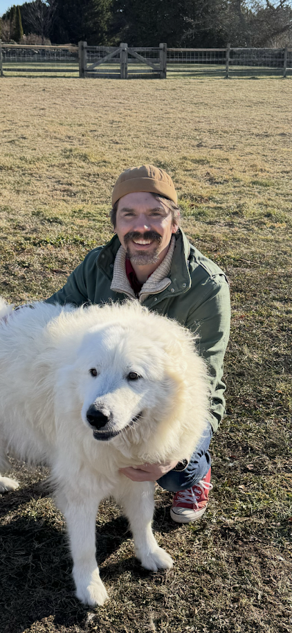 A man smiling and kneeling outdoors on a grassy field, holding a large fluffy white dog with a thick coat.