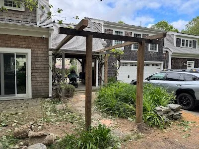 A wooden pergola frame in a residential front yard with plants and rocks.