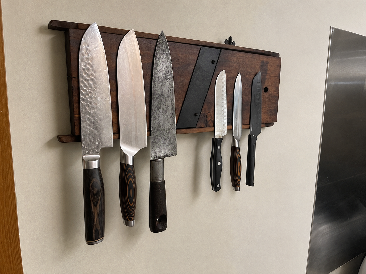 Six kitchen knives with various blade styles hanging on a magnetic strip on a beige wall.