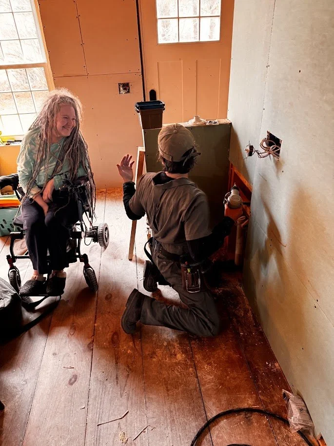 A woman in a wheelchair and a man kneeling on a wooden floor inside a room under construction, with exposed wiring and an unfinished wall. The woman is smiling and waving, the man is working on outlets or wiring.
