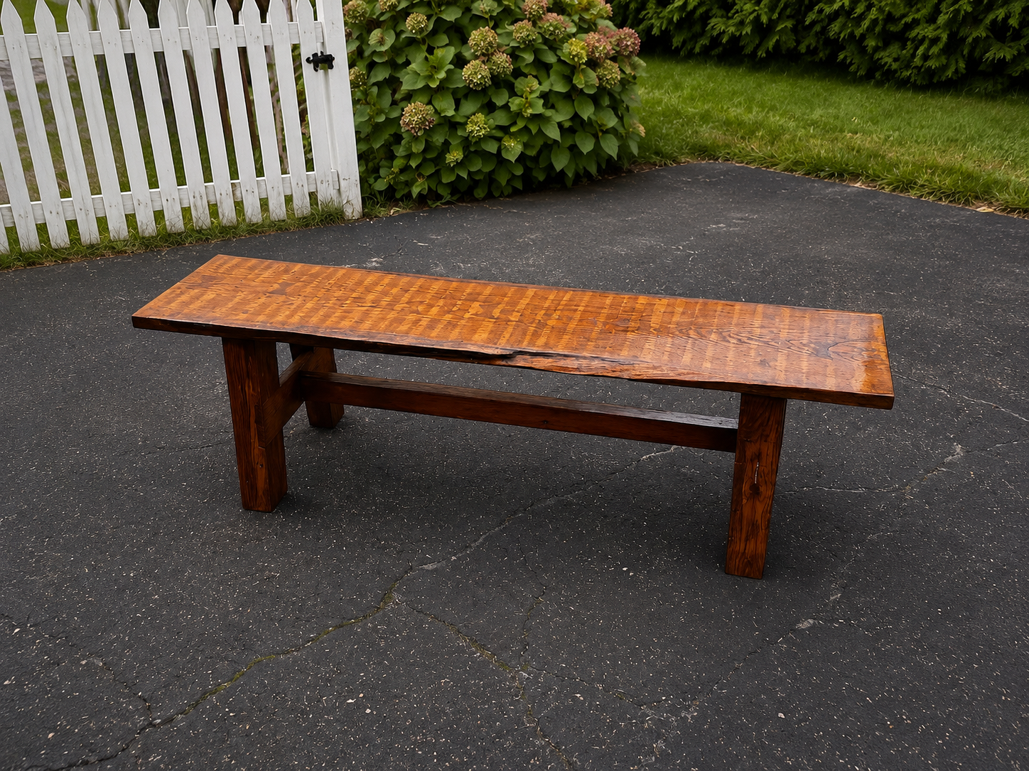 A wooden outdoor bench on a cracked asphalt driveway with green bushes and a white picket fence in the background.
