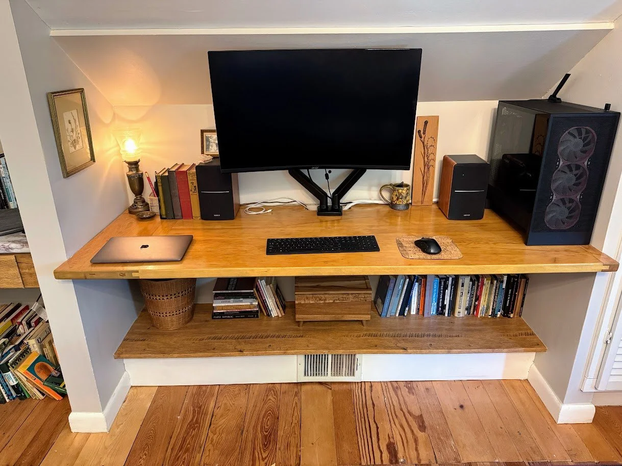 A home office desk made of wood with a bookshelf below. The desk holds a closed laptop, a keyboard, a mouse, and multiple speakers. Behind the monitor are books and a small decorative painting. To the right, there is a computer tower with fans visible on the side. On the left, a table lamp with a glowing bulb, books, and decorative items. The wall above the desk features framed artwork, and the desk is situated in a cozy room with wooden flooring.