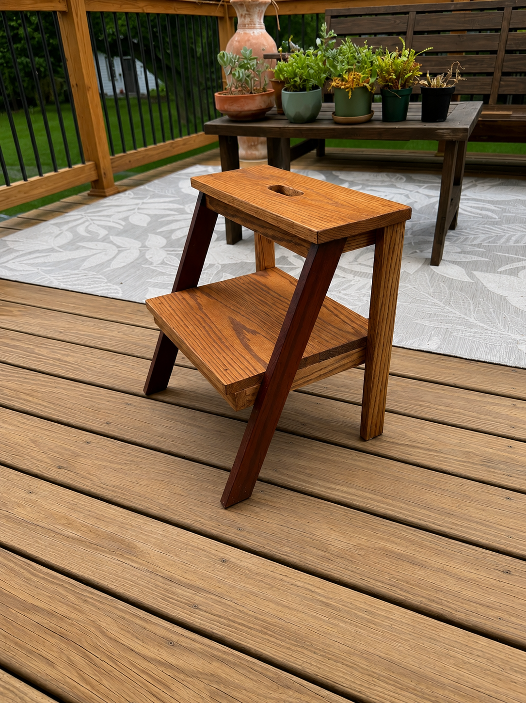 A wooden outdoor deck with a small wooden step stool in front of a black table holding potted plants and a pink vase, with a white patterned rug partially under the table.