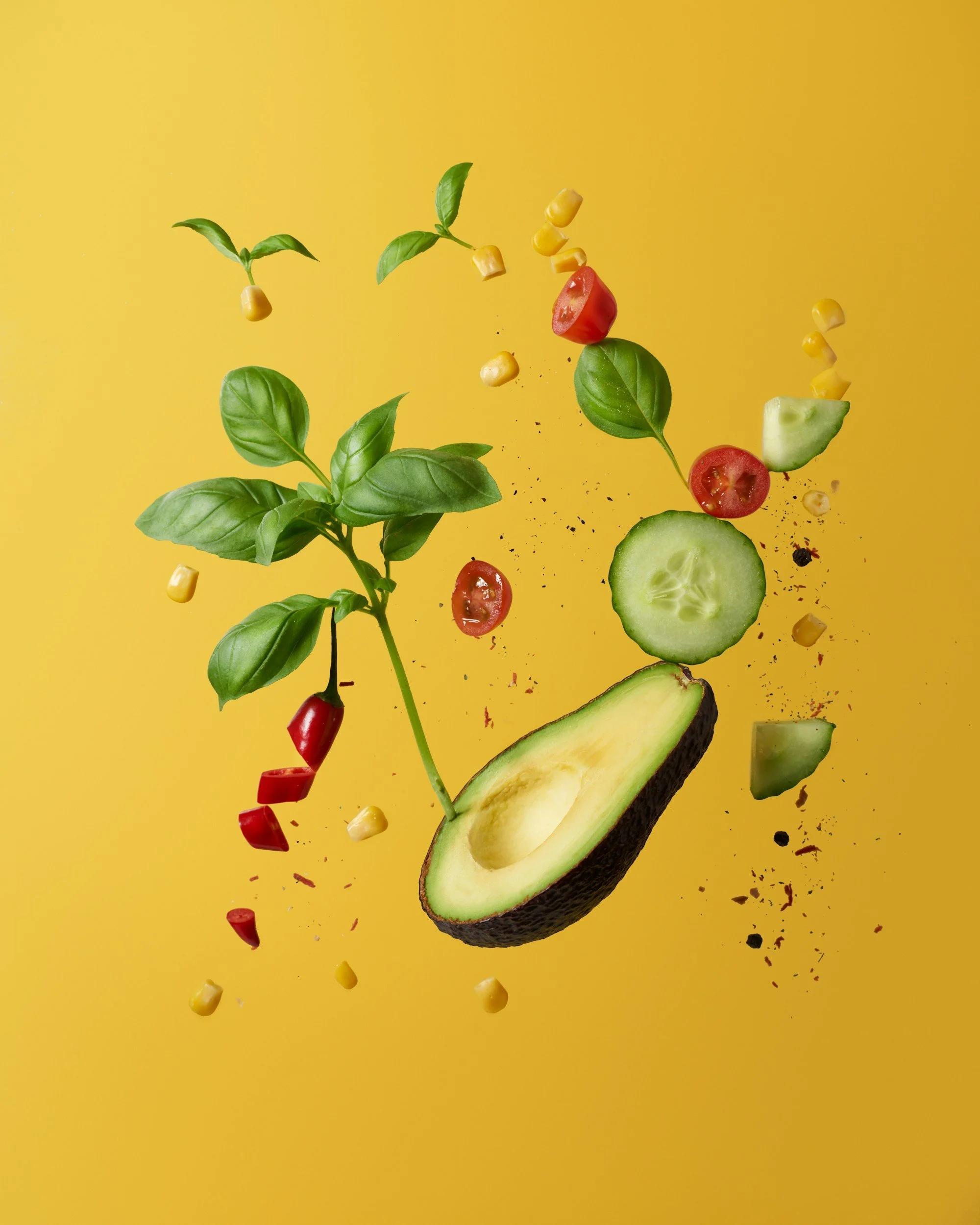 Various fresh vegetables and herbs, including cherry tomatoes, slices of cucumber, avocado, corn, and basil, suspended in mid-air against a yellow background.