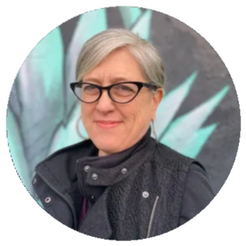 A woman with short gray hair and black glasses smiling, wearing a black vest over a dark shirt, standing outdoors with green and blue abstract background.