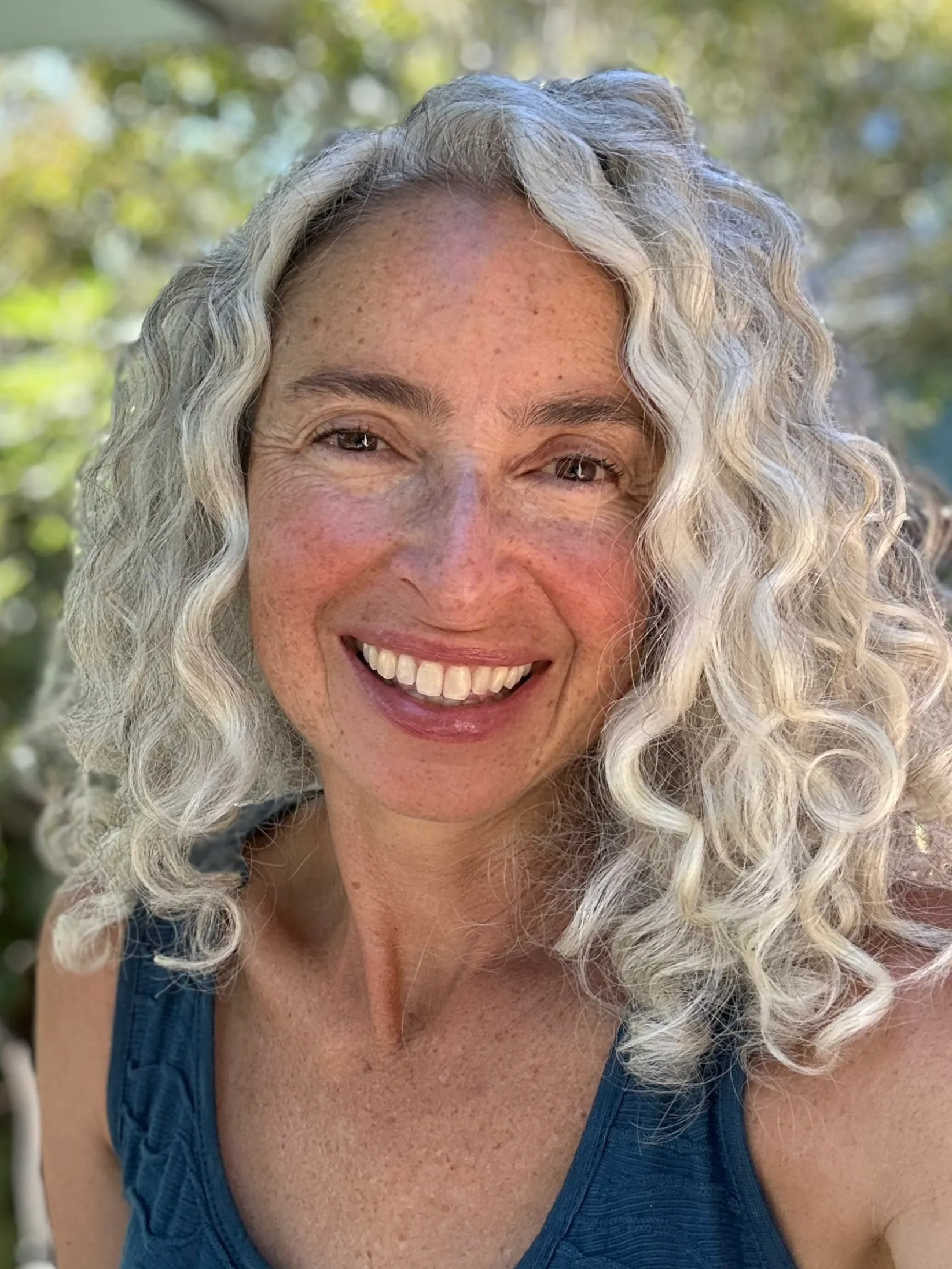Close-up of a smiling woman with curly gray hair and freckles outdoors with blurred greenery in the background.