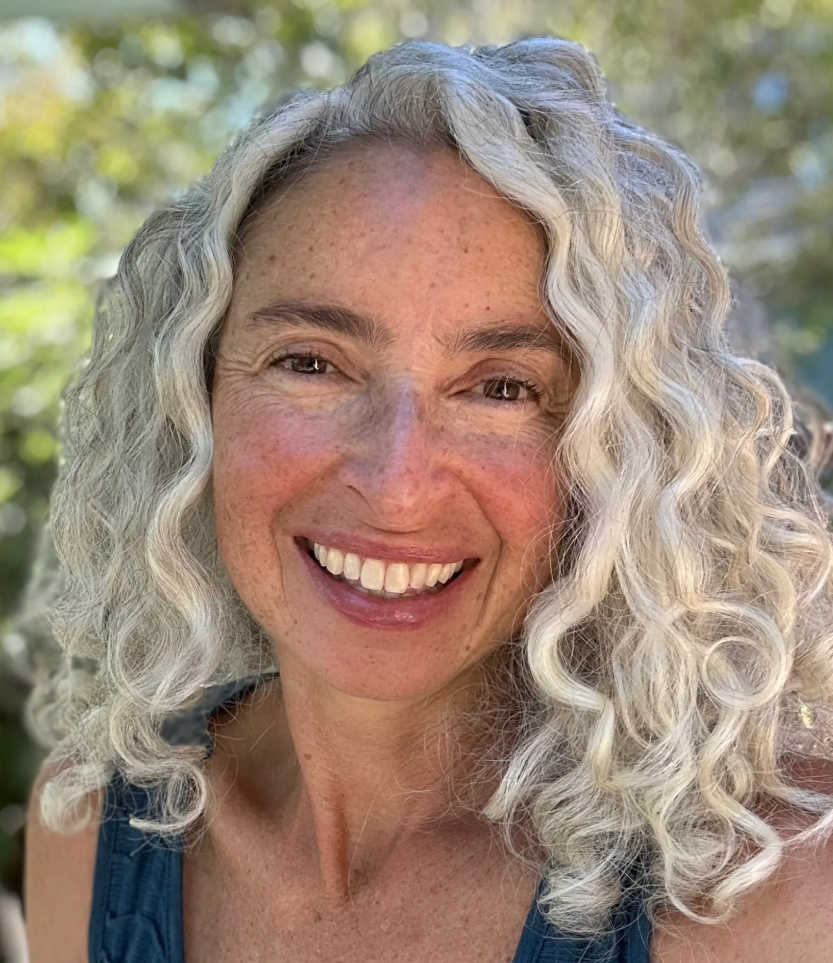 Close-up of a smiling woman with long, curly gray hair, freckles, and light brown eyes, outdoors with a blurred background of trees.