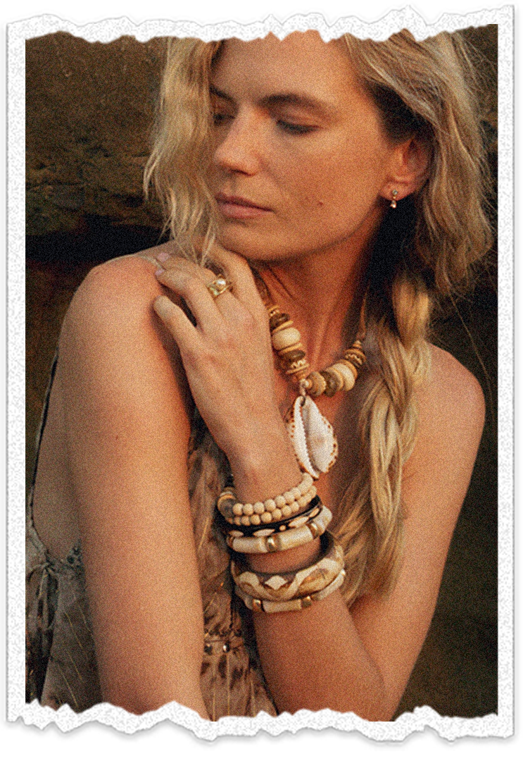 A woman with blonde, wavy hair styled in a side braid, wearing a sleeveless top, multiple beaded and shell jewelry, and earrings, resting her hand on her shoulder with a serene expression.