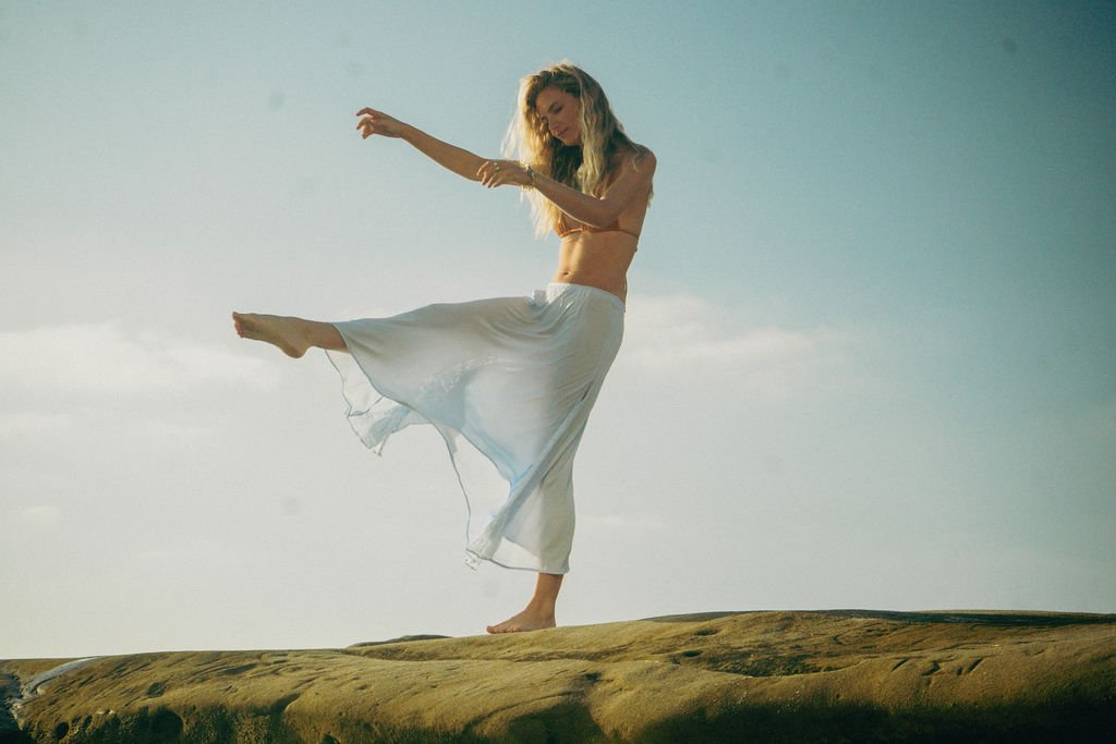 A woman in a white flowing skirt and top dancing on a rock formation outdoors with a cloudy sky in the background.