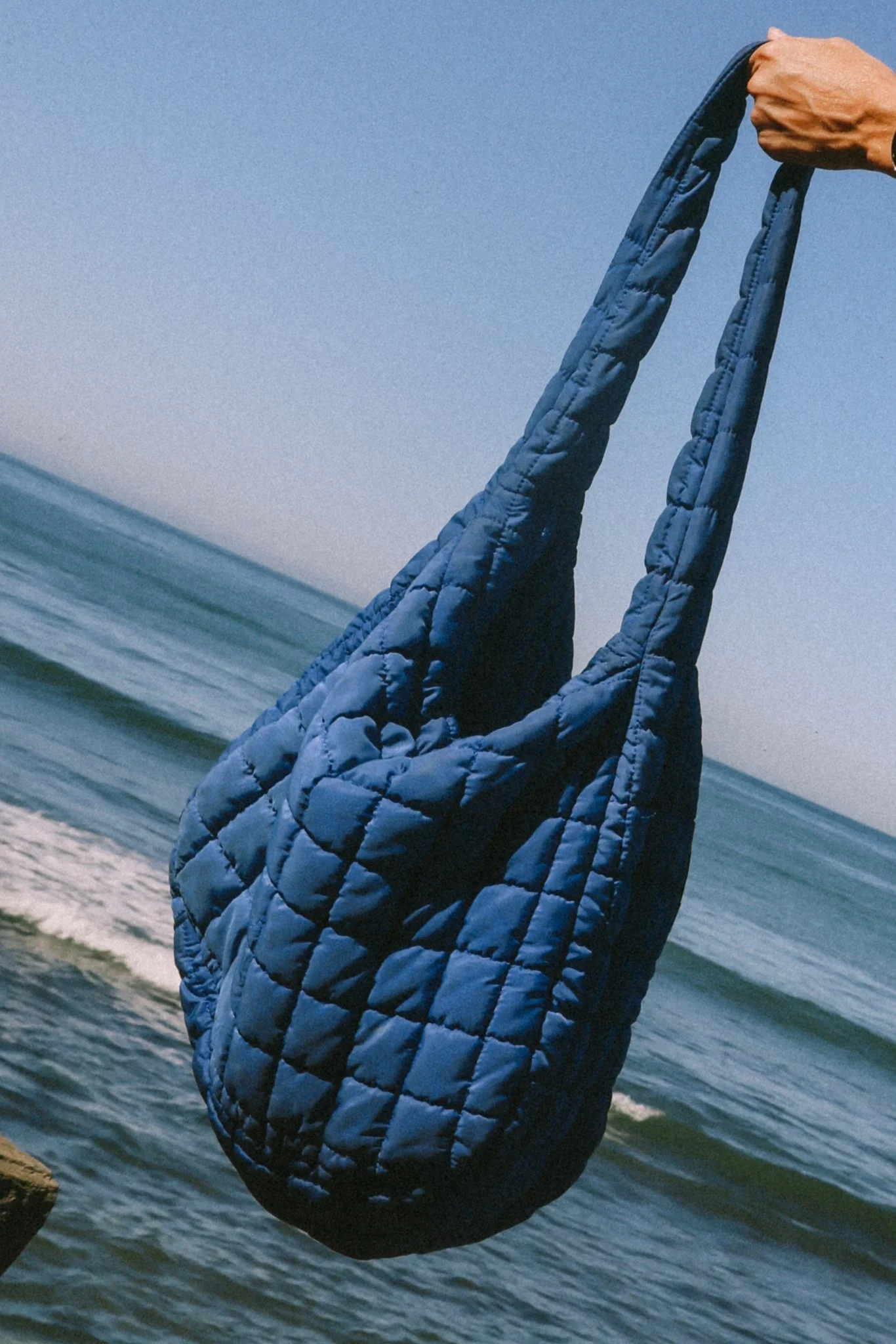 A person holding a blue quilted puffer jacket extended over the ocean shoreline with small waves and rocks, under a clear blue sky.