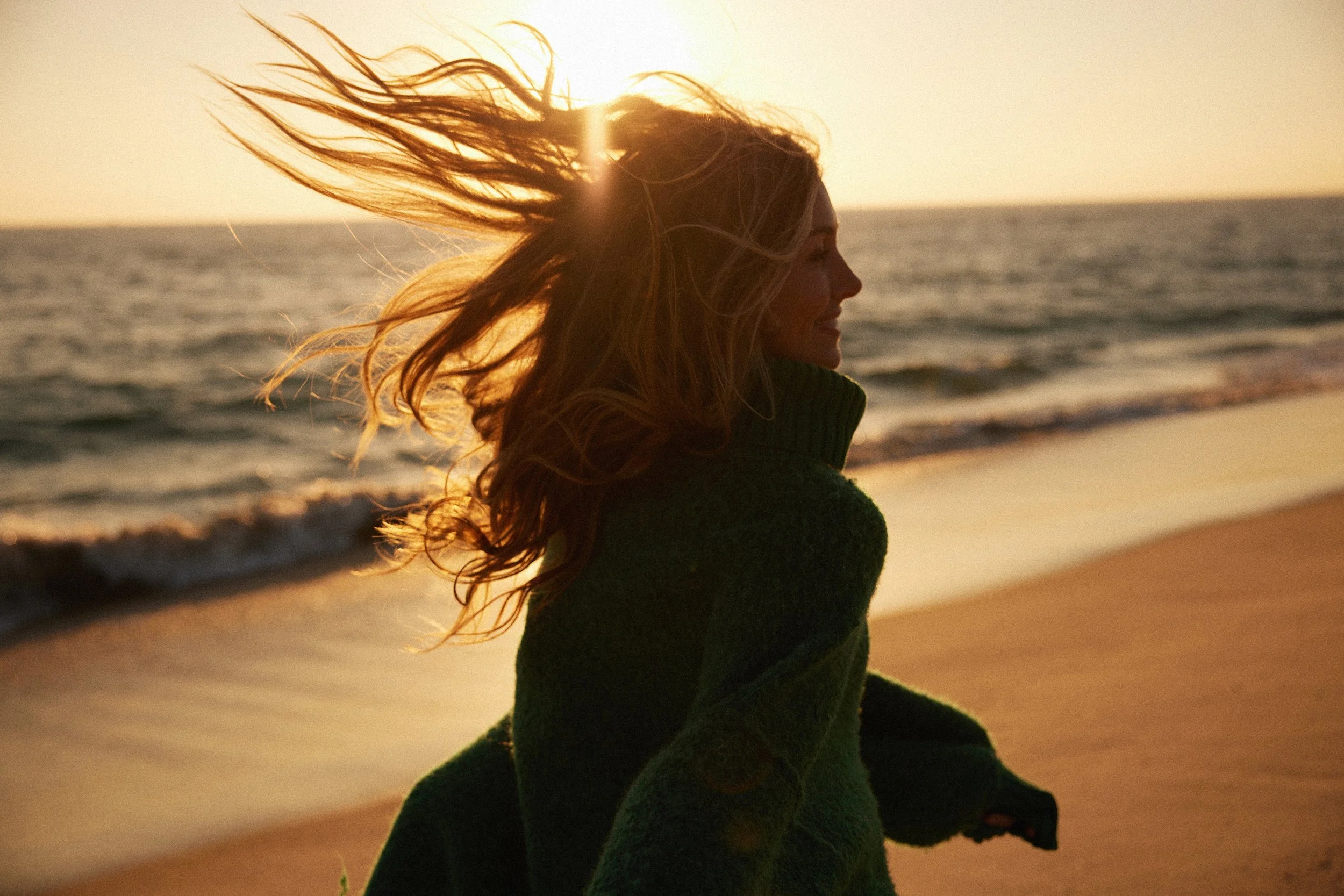 A woman with long hair standing on the beach at sunset, facing sideways with her hair blowing in the wind.