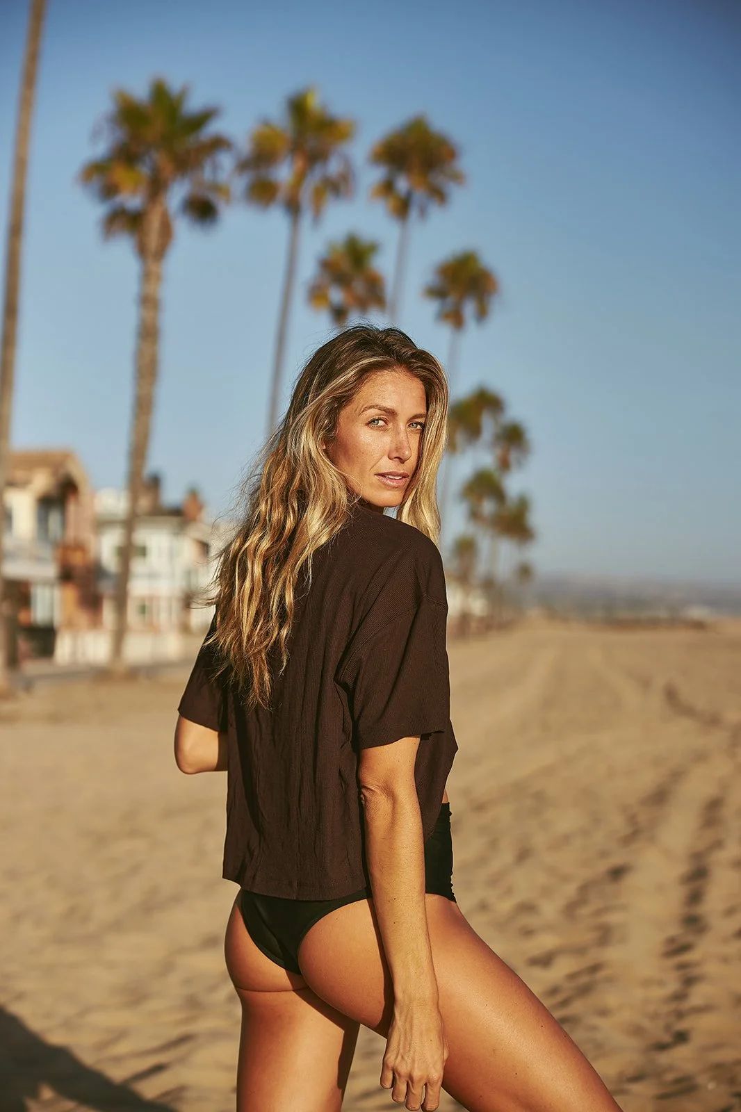 A woman standing on a sandy beach with palm trees in the background, wearing a black shirt and black bikini bottoms, looking over her shoulder at the camera during sunset.