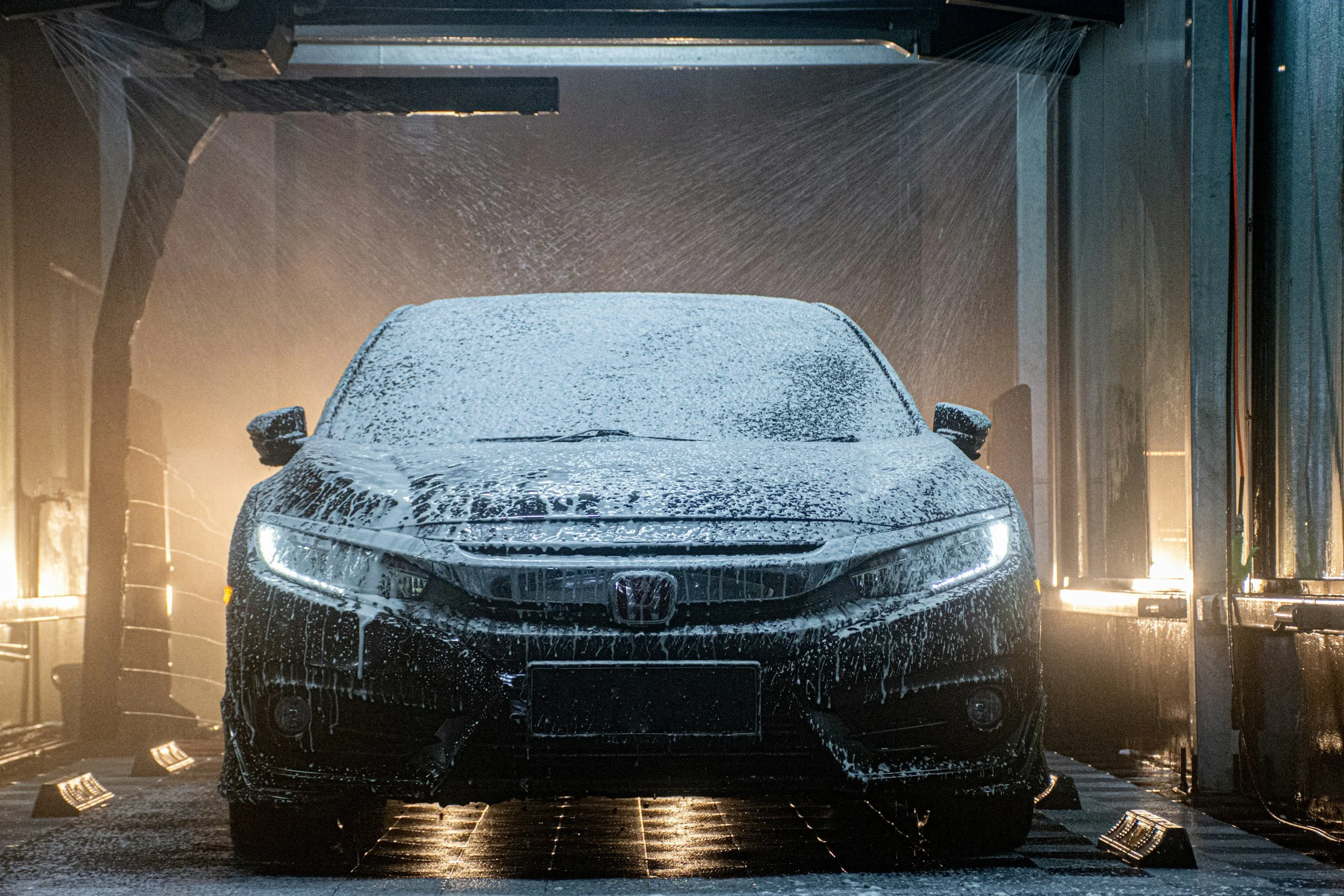 A black Honda car being washed in an automated car wash with soap and water, illuminated by bright lights, with water spray and soap suds covering the vehicle.