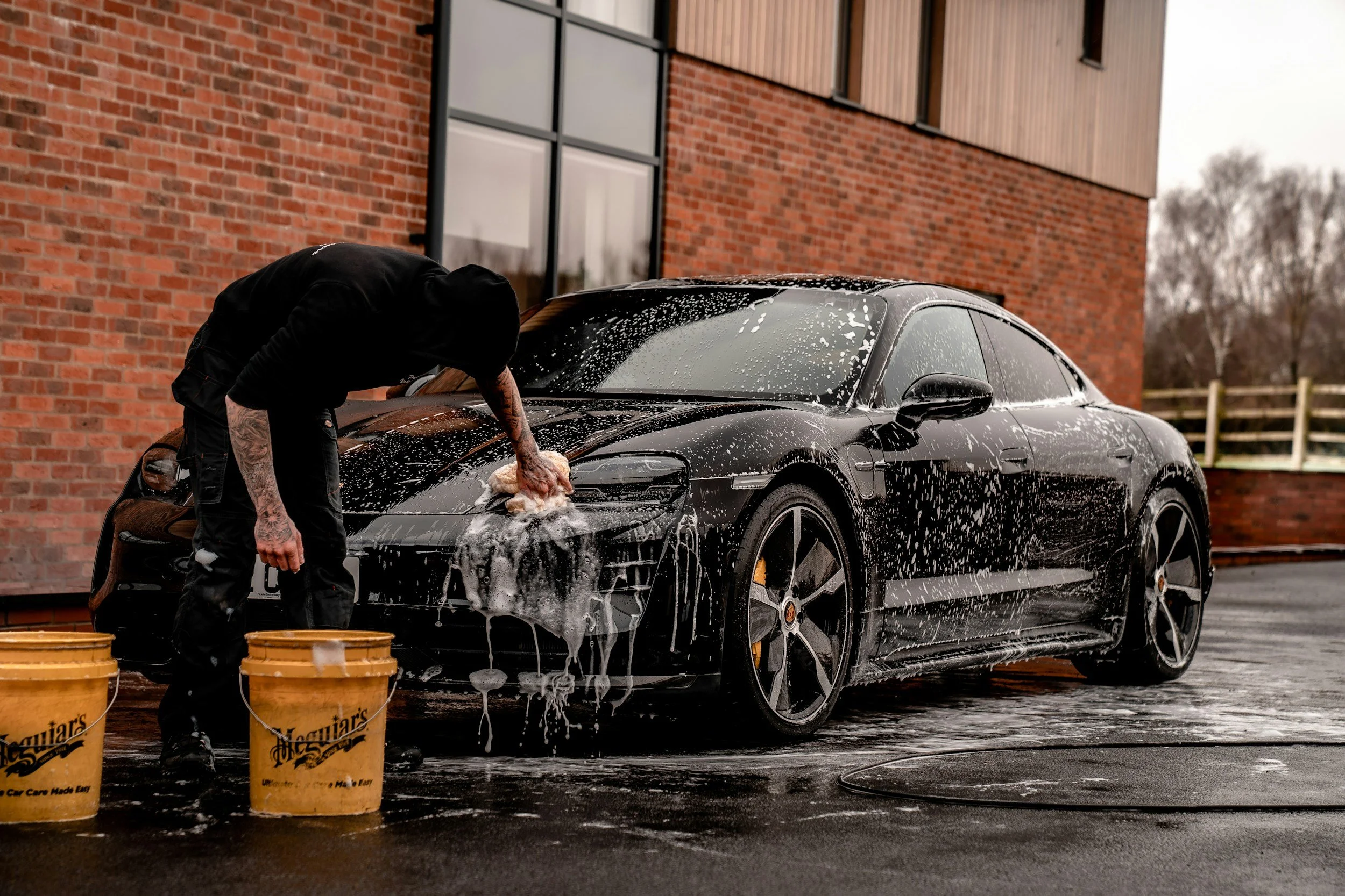Person washing a black sports car with soap and sponge outside near a brick building.