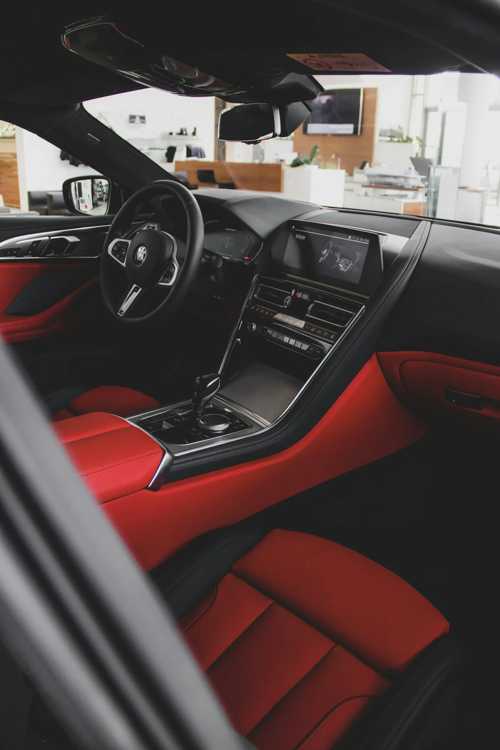 Interior view of a luxury car showing a red and black leather seat, steering wheel, dashboard with digital displays, and a modern infotainment system. Outside features a showroom with polished floors and bright lighting.