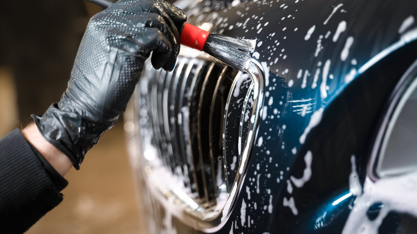 A person wearing black gloves cleaning the front grille of a black car with a brush and soap.