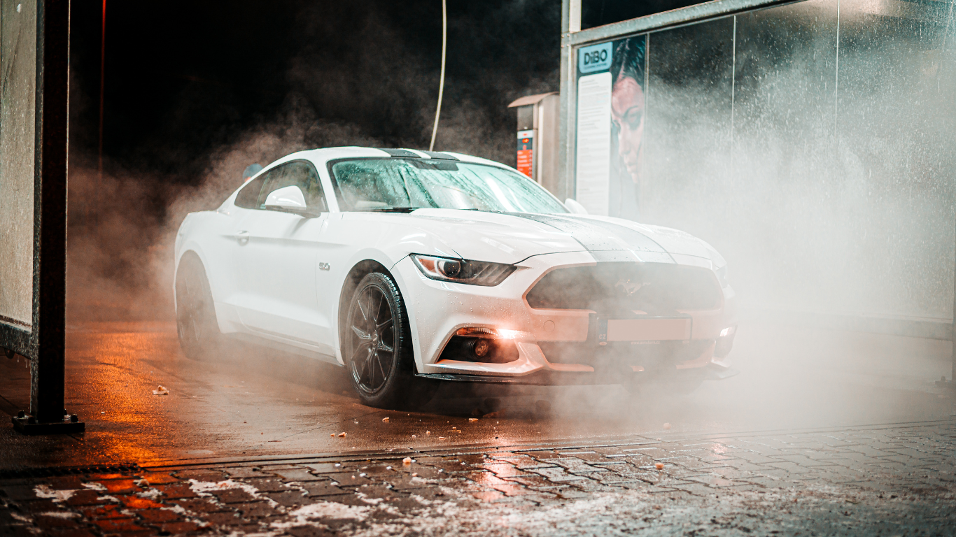 A white Ford Mustang during a car wash, with water spray and steam surrounding it.