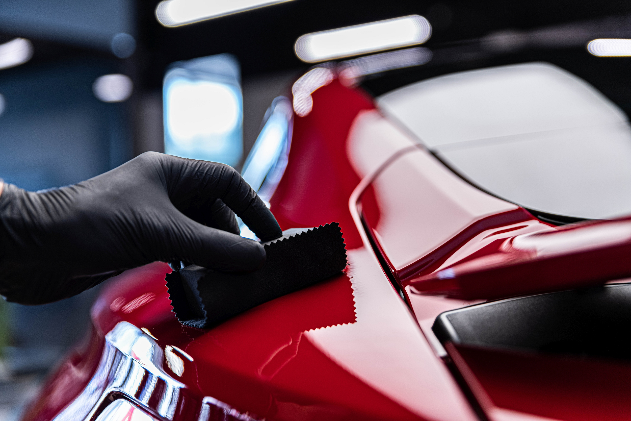 A person in black gloves holding a microfiber cloth polishing a red car's surface in a garage or workshop.
