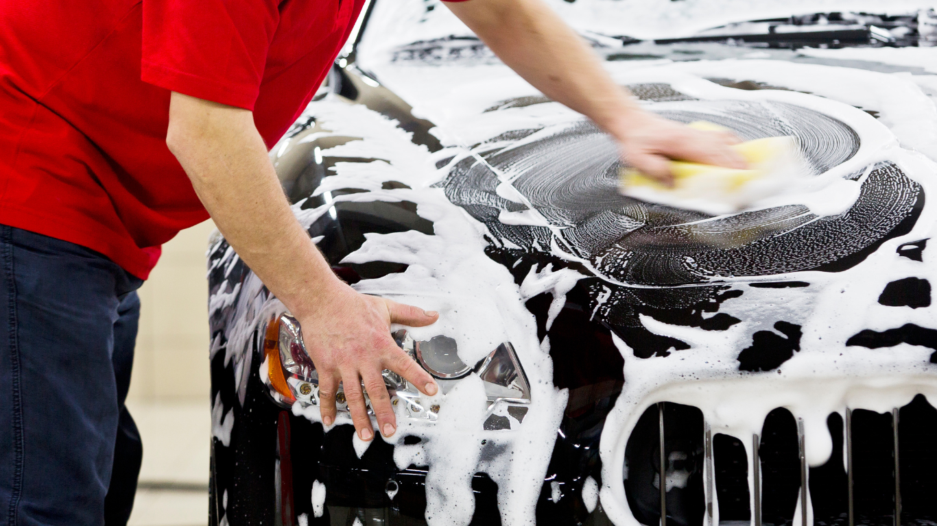 Person washing a black car with soap and a sponge in a garage.