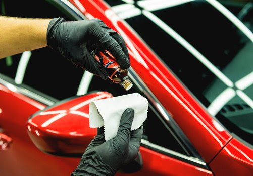A person wearing black gloves holding a small container of polishing compound and a white cloth, polishing a red car's headlight.