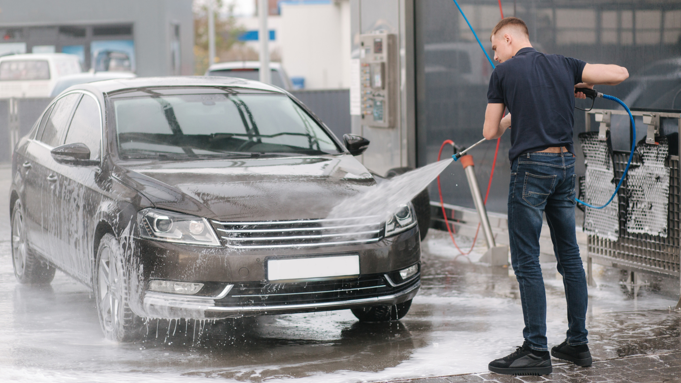 Man washing a black sedan car with a high-pressure water spray at a car wash facility.