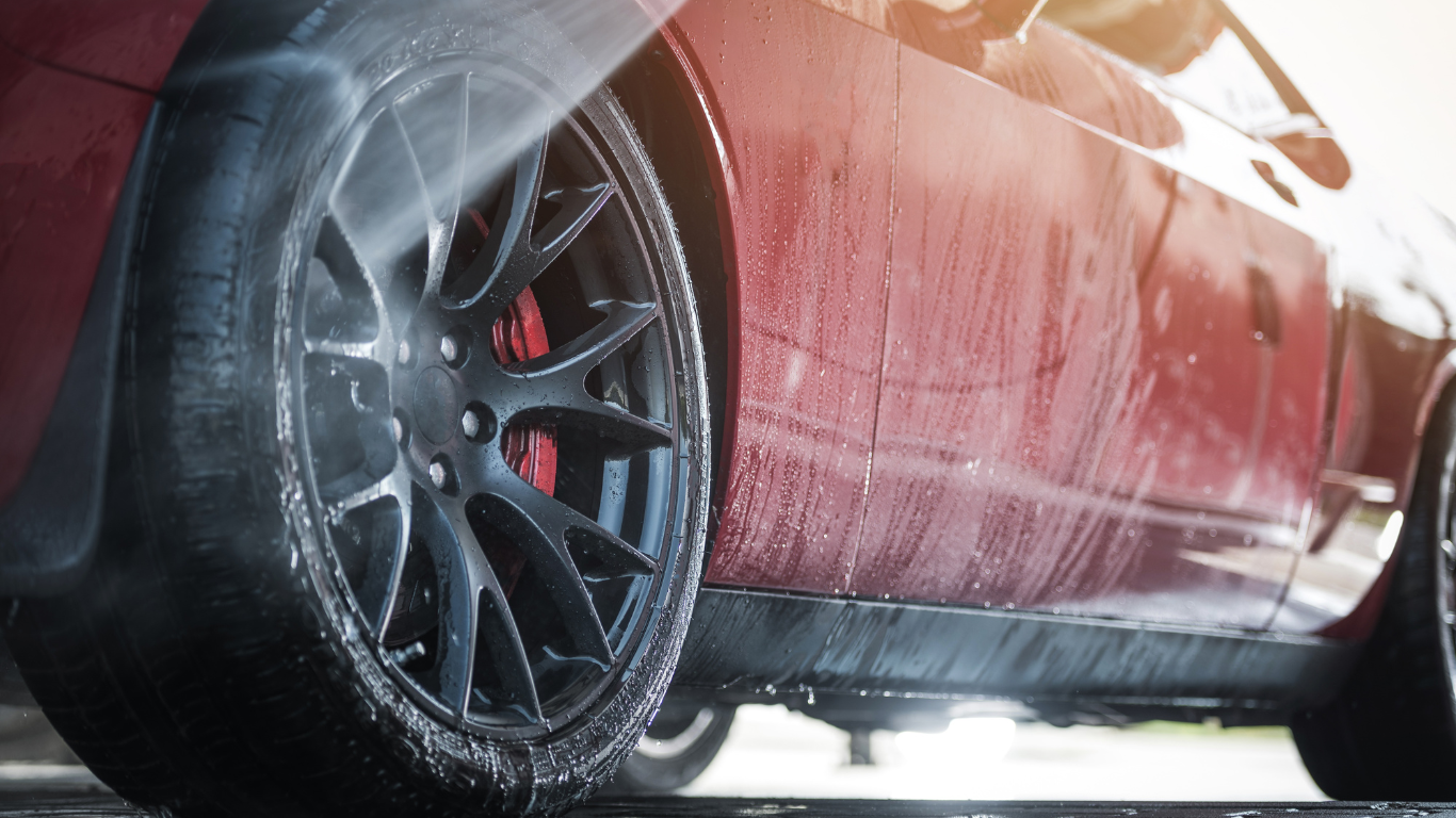 Red sports car being washed with water and soap, focusing on the rear wheel and side panel.