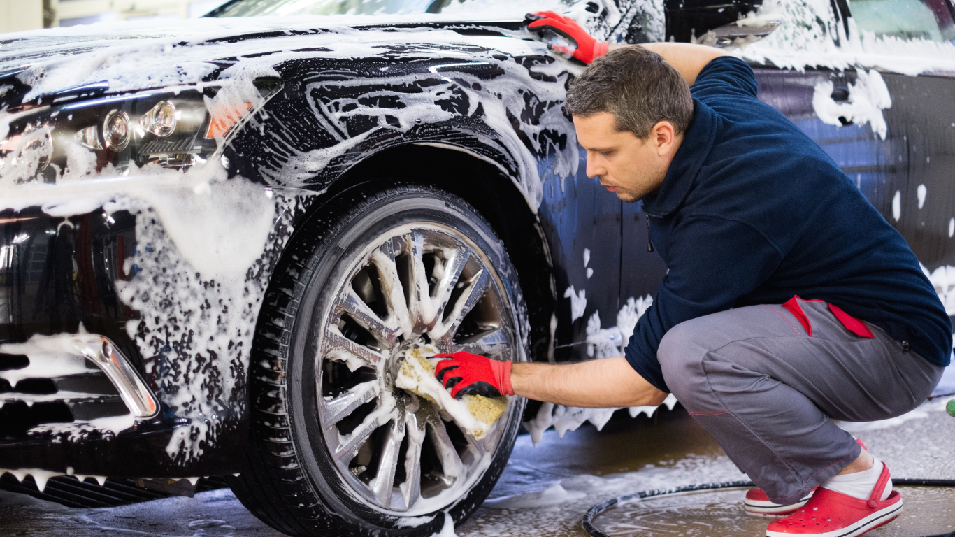A man cleaning a black car with soap and a sponge, wearing red gloves and red Crocs.