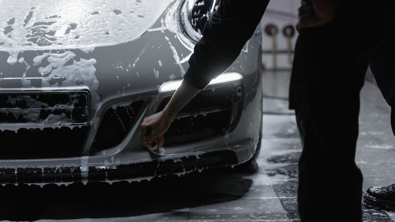 Person washing a silver car in a car wash, with soap suds covering the front of the vehicle.