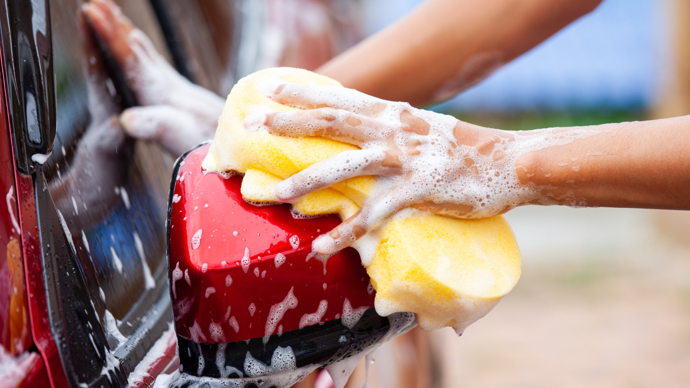 Person washing the side mirror and door of a red car with a yellow sponge and soap suds.