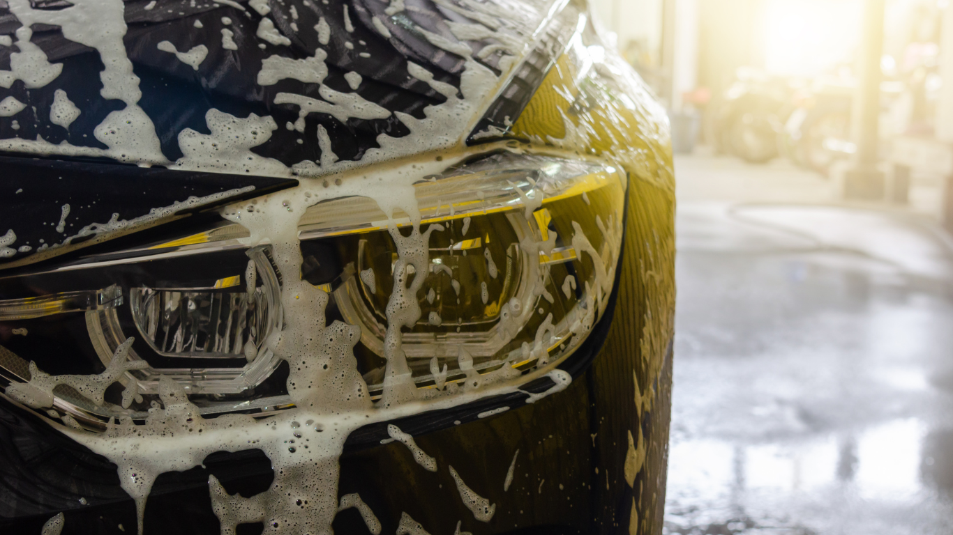 Close-up of a black car's front headlight covered in soap suds during a car wash, with a wet surface and sunlight in the background.