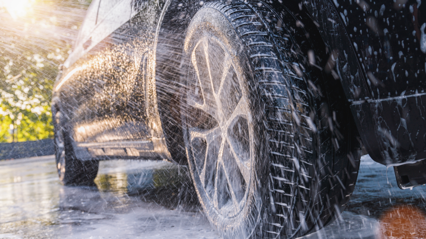 Close-up of a black car spinning in a car wash, with water spraying off the tires and wheel, sunlight shining on the wet surface.